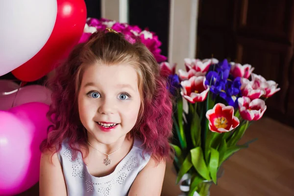 Pequena menina bonita com cabelo encaracolado rosa em um vestido de prata em tulipas vermelhas e com bolas multi-coloridas — Fotografia de Stock