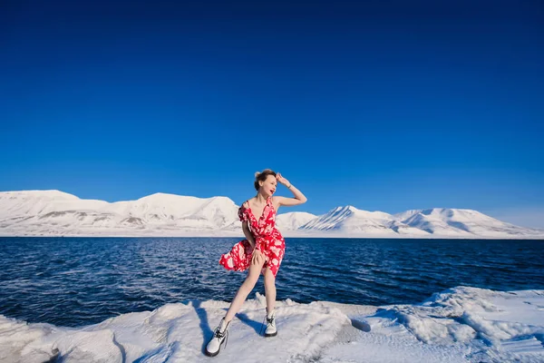 Chica delgada hermosa rubia en un vestido rojo en la helada sobre un fondo de montañas svalbard en la ciudad de Spitsbergen Longyearbyen, en tiempo soleado — Foto de Stock