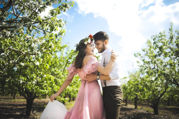 少女と男の結婚式をお探しのカップルのドレス、花の花輪と背景庭園、青空の彼女の頭の上飛ぶピンクのドレスと彼らの抱擁 — ストック写真