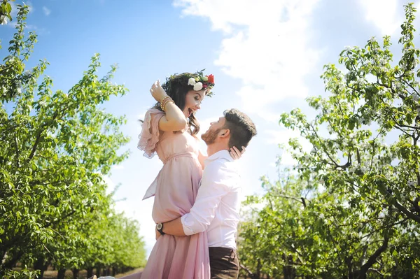Couple d'une fille et un gars à la recherche d'une robe de mariée, une robe rose volant avec une couronne de fleurs sur la tête sur un jardin d'arrière-plan et le ciel bleu, et ils étreignent — Photo