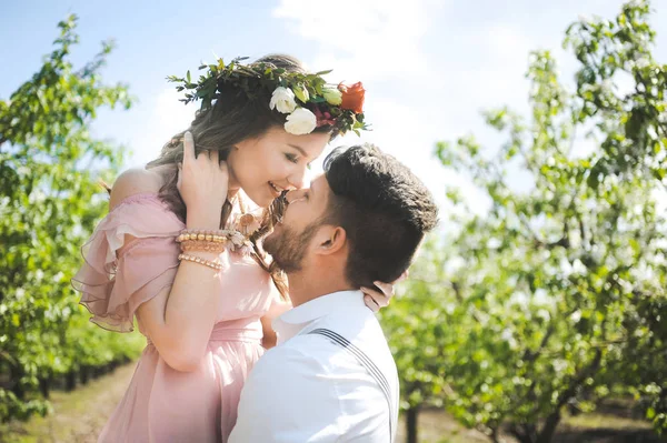 Pareja de una chica y un chico en busca de un vestido de novia, un vestido rosa volando con una corona de flores en la cabeza en un jardín de fondo y el cielo azul, y se abrazan — Foto de Stock