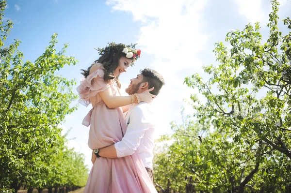Paar von einem Mädchen und einem Mann auf der Suche nach einem Hochzeitskleid, einem rosa Kleid, das mit einem Blumenkranz auf dem Kopf vor einem Garten im Hintergrund und dem blauen Himmel fliegt, und sie umarmen sich — Stockfoto