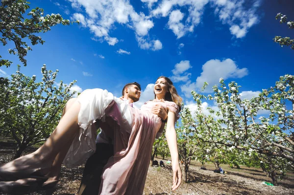 Пара девушка и парень ищет свадебное платье, розовое платье летит с венком цветов на голове на заднем плане сада и голубое небо, и они обнимаются — стоковое фото