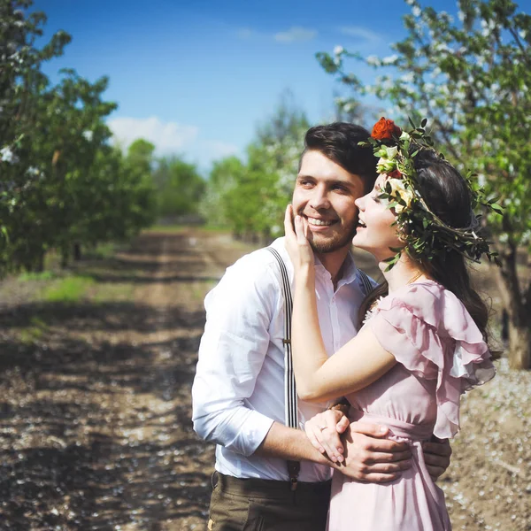 Pareja de una chica y un chico en busca de un vestido de novia, un vestido rosa volando con una corona de flores en la cabeza en un jardín de fondo y el cielo azul, y se abrazan — Foto de Stock