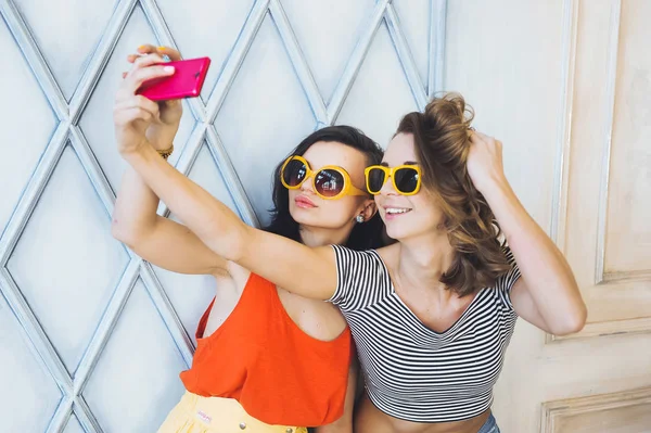 Krásný mladý pár módní holky blondýnka a brunetka v zářivě žluté šaty a sluneční brýle, pózování a usmívá se na kameru a dělá smartphone selfi — Stock fotografie