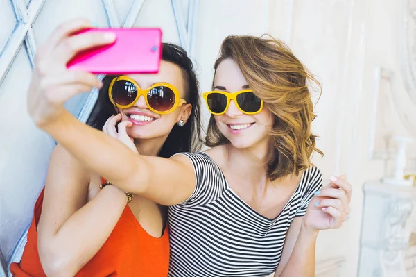 Krásný mladý pár módní holky blondýnka a brunetka v zářivě žluté šaty a sluneční brýle, pózování a usmívá se na kameru a dělá smartphone selfi — Stock fotografie