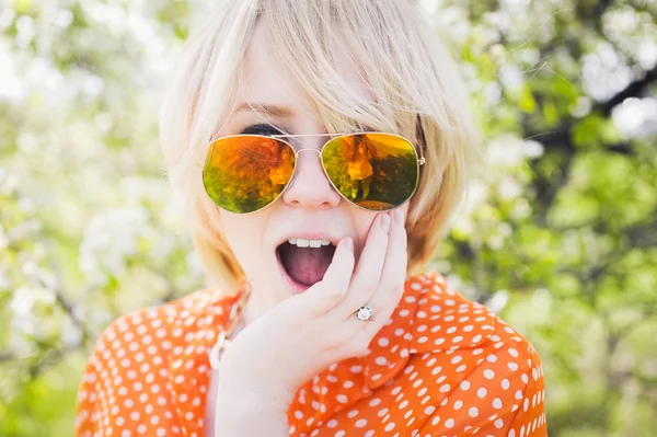 Portret buitenshuis jong mooi meisje in een oranje hipster blonde vrolijke heldere blouse en glimlachend in zonnebril op een achtergrond bloeiende bomen met witte tanden — Stockfoto