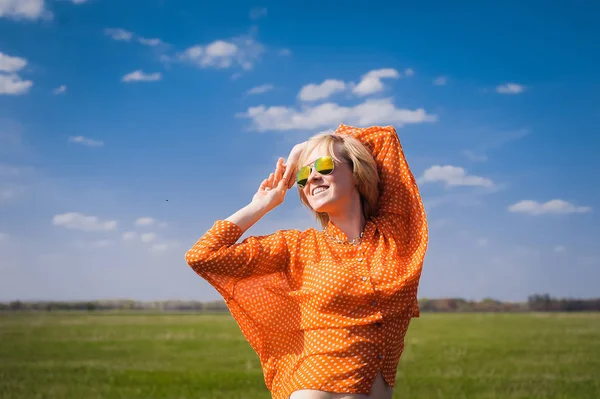 Retrato ao ar livre jovem menina bonita em uma blusa de ponto alegre brilhante laranja hipster loira e sorrindo em óculos de sol em um fundo árvores florescentes com dentes brancos — Fotografia de Stock