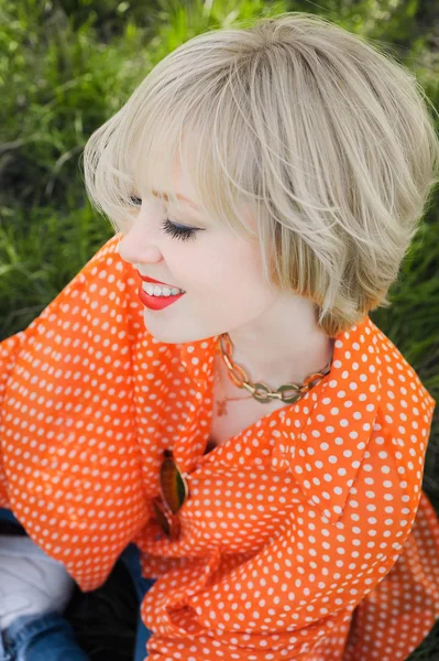 Retrato ao ar livre jovem menina bonita em uma blusa de ponto alegre brilhante laranja hipster loira e sorrindo em óculos de sol em um fundo árvores florescentes com dentes brancos — Fotografia de Stock