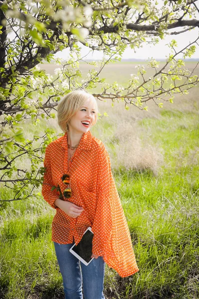 Portrait outdoors young beautiful girl in an orange hipster blonde bright cheerful  dot blouse , smiling red plump lips on the background of green grass with laptop — Stock Photo, Image