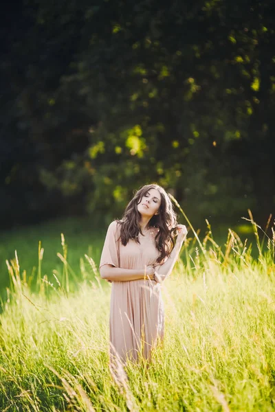 Beautiful attractive lady of a young slender brunette girl with long dress walking through a green park with flowers at sunset lifestyle — Stock Photo, Image