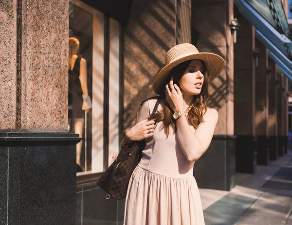 Retrato de una delgada joven blogger hermosa morena en el centro de Dusseldorf con un vestido de pastel y un sombrero de señora con gafas de sol caminando poses y sonriendo en la puesta de sol — Foto de Stock