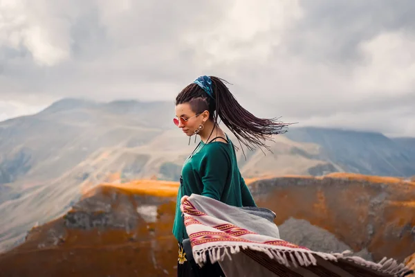 Porträtt av en ung vacker hipster flicka i solglasögon med hår dreadlock i en boho gypsy hippie stil kläder på en bakgrund av molnig himmel och hösten bergen i Georgien Dans — Stockfoto