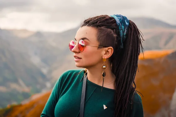 Retrato de uma jovem linda garota hipster em óculos de sol com temor de cabelo em um boho roupas estilo hippie cigano em um fundo de céu nublado e montanhas de outono da Geórgia dançando — Fotografia de Stock