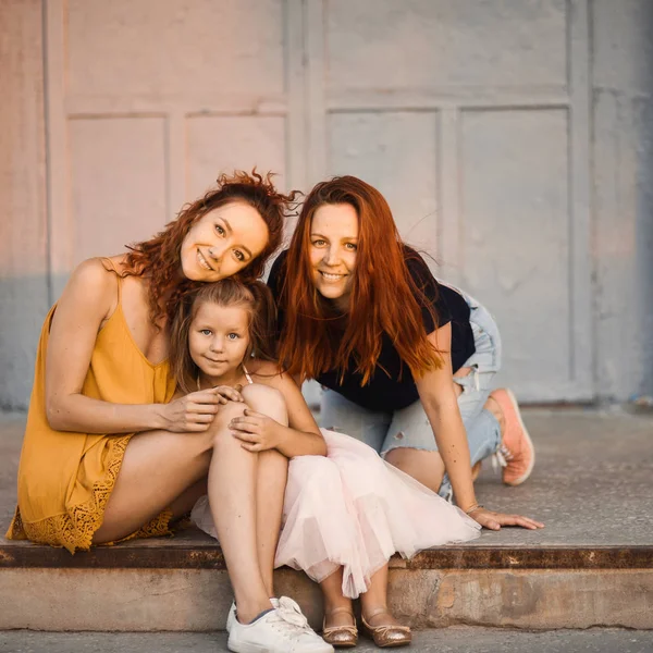 young beautiful red-haired mother and sister with a cute daughter smiling embraced on a sunny day, family portrait