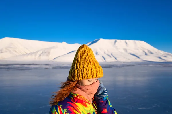 一个女孩的特写在彩虹羽绒服黄色针织帽与红色头发美丽的年轻头发在山的背景下朗伊尔城挪威天北极冬季 — 图库照片