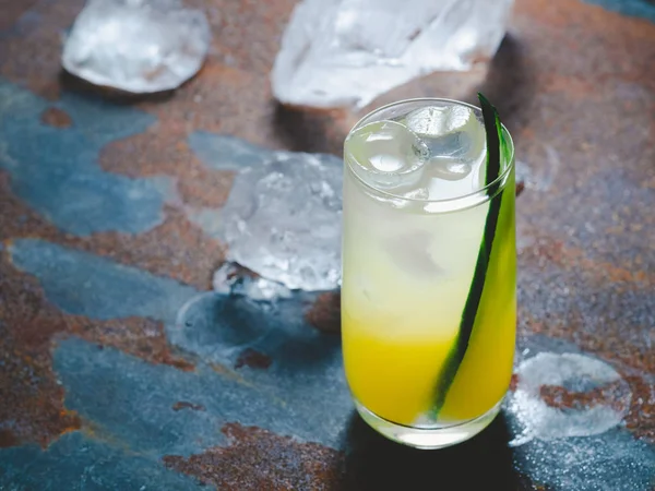 Refreshing cold yellow and white cocktail with the piece of cucumber and ice in a tall transparent grass, rusty metal background. Alcoholic or non-alcoholic drink. horizontal orientation