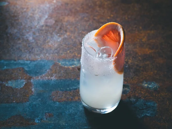 Coquetel roily branco com cubos de gelo em um vidro transparente liso decorado com pedaço de toranja fresca à direita em um fundo enferrujado. Luz de fundo, orientação vertical, espaço de texto — Fotografia de Stock