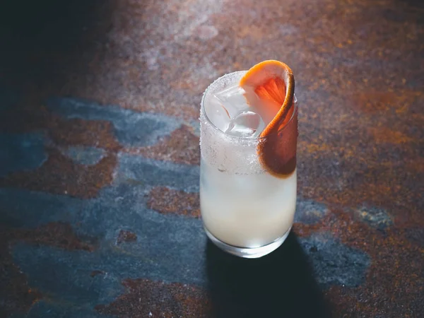 Vit roily cocktail med isbitar i en smidig genomskinligt glas inredda med bit färsk grapefrukt till höger på en rostig bakgrund. Bakgrundsbelysning, lodrät orientering, text utrymme — Stockfoto