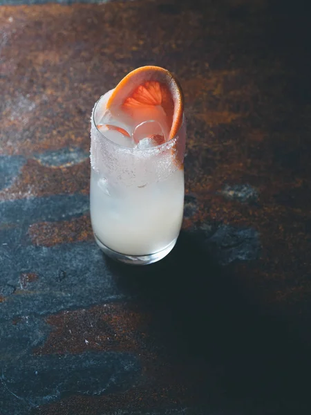 Vit roily cocktail med isbitar i en smidig genomskinligt glas inredda med bit färsk grapefrukt till höger på en rostig bakgrund. Bakgrundsbelysning, lodrät orientering, text utrymme — Stockfoto