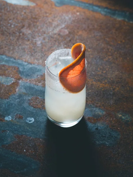 Vit roily cocktail med isbitar i en smidig genomskinligt glas inredda med bit färsk grapefrukt till höger på en rostig bakgrund. Bakgrundsbelysning, lodrät orientering, — Stockfoto