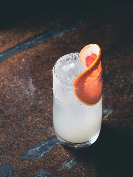 Vit roily cocktail med isbitar i en smidig genomskinligt glas inredda med bit färsk grapefrukt till höger på en rostig bakgrund. Lynnig ljus, vertikal orientering — Stockfoto