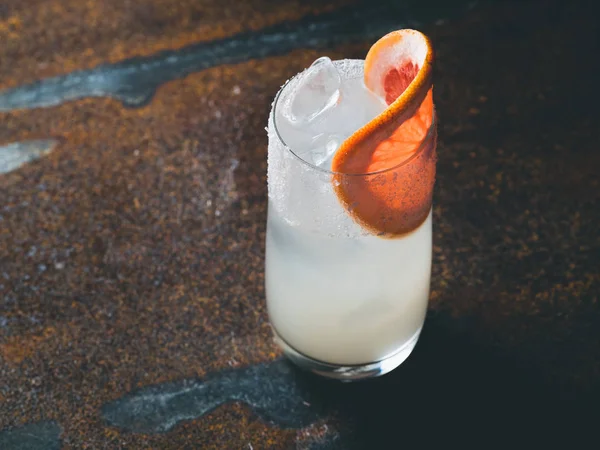 Vit roily cocktail med isbitar i en smidig genomskinligt glas inredda med bit färsk grapefrukt till höger på en rostig bakgrund. Bakgrundsbelysning, horisontell orientering, text utrymme — Stockfoto