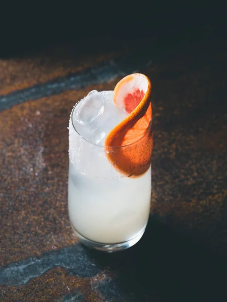 White roily cocktail with ice cubes in a smooth transparent glass decorated with piece of fresh grapefruit on the right on a rusty background. Moody light, horizontal orientation — Stock Photo, Image
