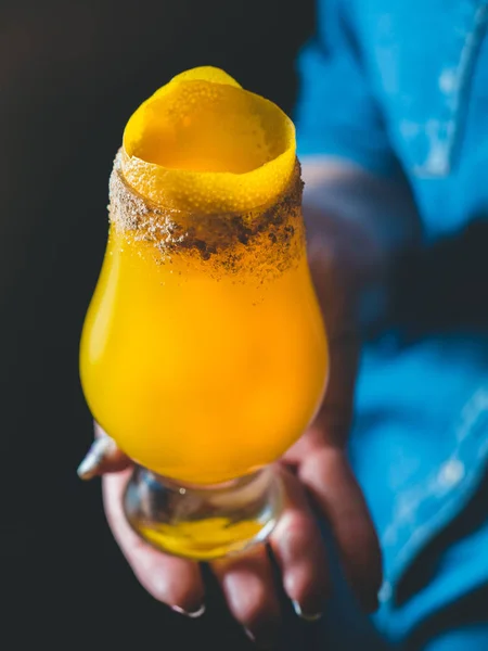 Coquetel de manga amarela com raspas de limão e crusta de açúcar em um copo de tulipa, mão feminina / barman segurando um copo com o coquetel, fundo de camisa de jeans azul. Coquetel close-up, orientação vertical — Fotografia de Stock