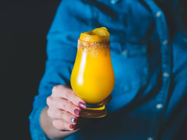 Коктейль из желтого манго с лимонной цедрой и сахарной корой в тюльпановом бокале, женская рука / бармен, держащие стакан с коктейлем, синие джинсы на заднем плане. Коктейль в центре — стоковое фото