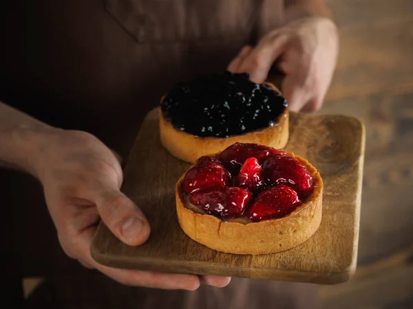 Stroberi tart dan blueberry tart pada papan kayu close-up. Tangan laki-laki memegang memotong papan dengan kue yang indah. Foto horisontal — Stok Foto