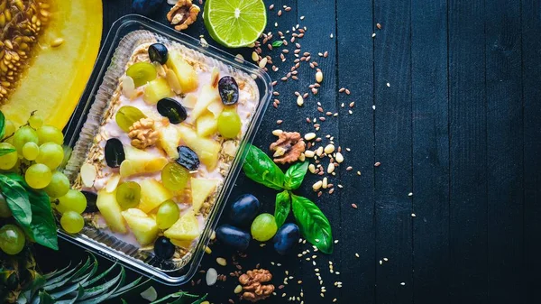 Porridges yoğurt, ananas, üzüm ve karpuz. Sağlıklı bir diyet yemek. Öğle Yemeği boks. Siyah ahşap arka plan üzerinde. Üstten Görünüm. Metin için boş alan.