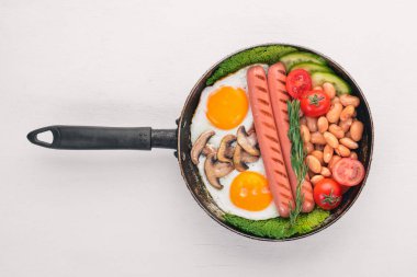 İngiliz kahvaltısı domates, sosis, fasulye, mantar, yumurta, tost ve otlar üstten görünüm. Beyaz ahşap zemin üzerinde.