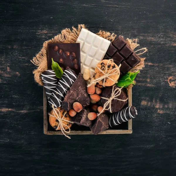 A set of milk chocolate and black chocolate in a wooden box with nuts and biscuits. On a black wooden background. Copy space for text.