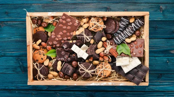 A set of milk chocolate and black chocolate in a wooden box with nuts and biscuits. On a blue wooden background. Copy space for text.