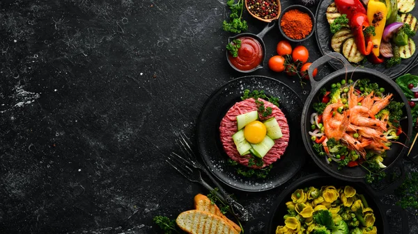Assorted food and dishes of vegetables, meat and fish on a black stone background. Top view. Free space for your text.
