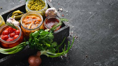 Sebze turşusu ve mantarlar siyah taş arka planda, tahta kutuda cam kavanozlarda. Üst görünüm.