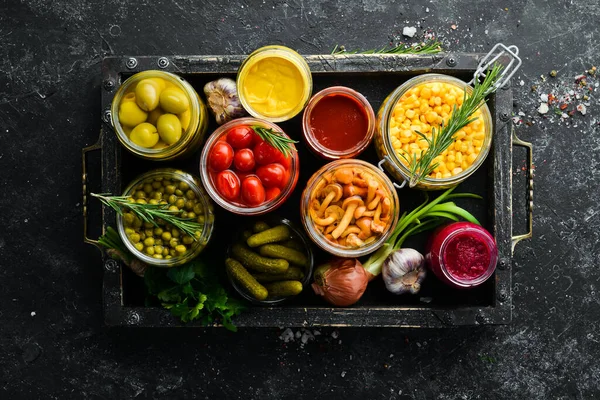 Sebze turşusu ve mantarlar siyah taş arka planda, tahta kutuda cam kavanozlarda. Üst görünüm.