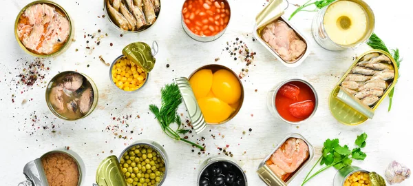 Food background in tin cans. Canned vegetables, beans, fish and fruits on a white wooden background. Top view.