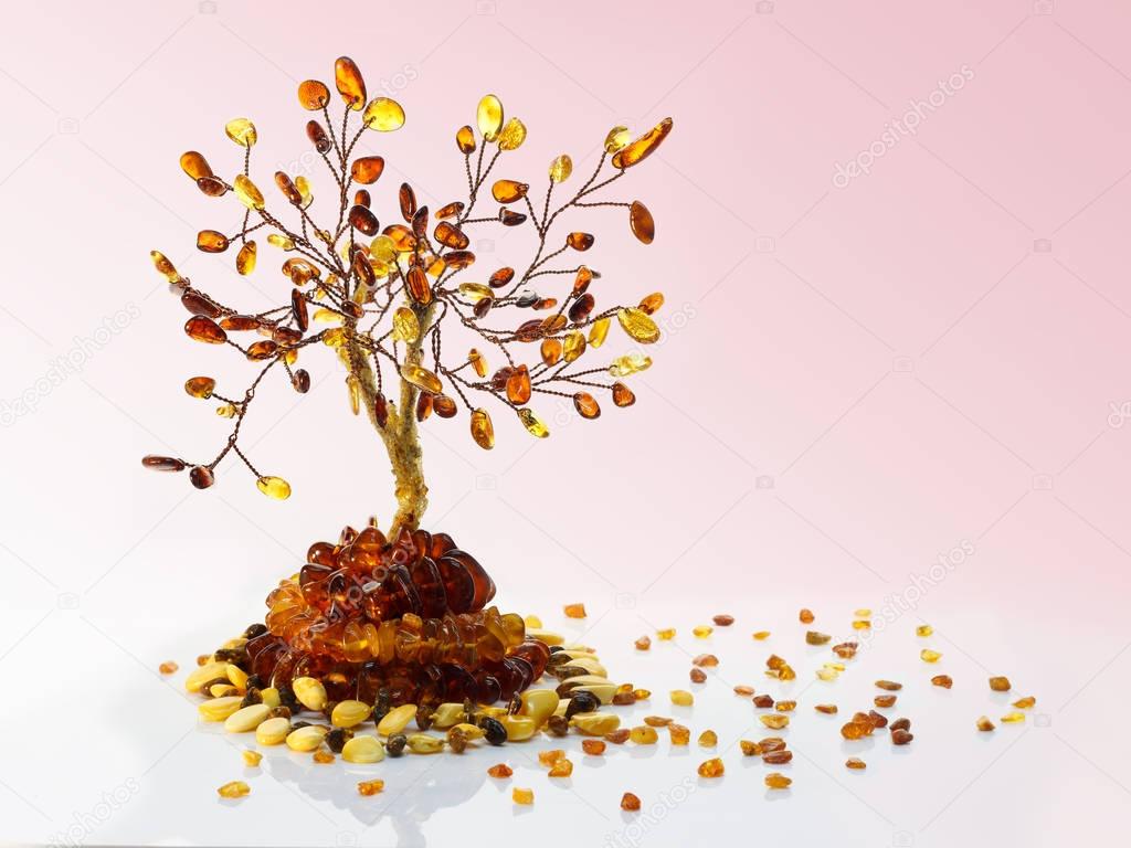 Decorative tree made withyellow and dark brown baltic amber, amber beads and small pieces of raw amber on white acrylic surface on pink gradient background