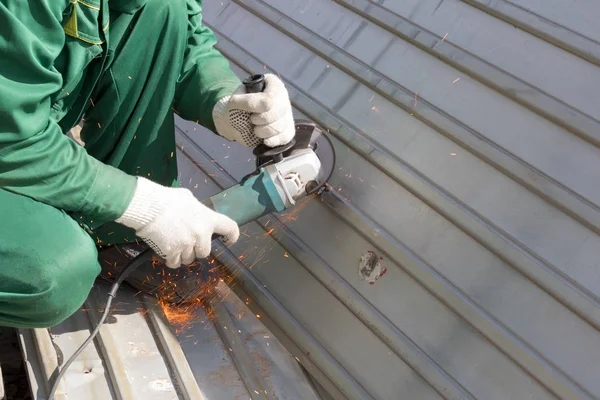 worker in green uniform and in rags gloves grinds corrugated steel structure for painting primer anti rust. Red sparks from  wheel.