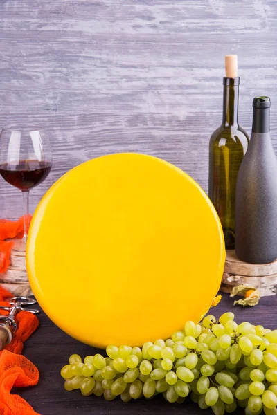 A round cheese in a yellow film with green grapes and a glass of wine