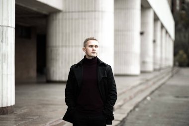 Siyah trençkotlu şık bir adam ve modernist bina geçmişinde polo yakalı.