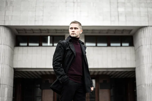 Stylish man in black trench coat and polo neck on modernist building background