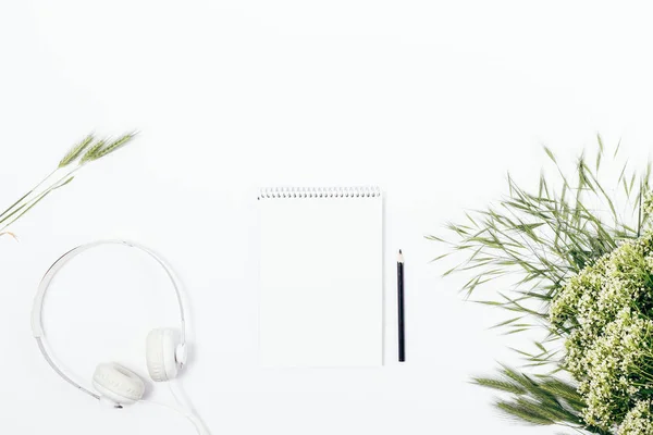 Blank notepad near bouquet of white wild summer flowers and headphones, top view. Pure feminine flat lay composition with copy space.