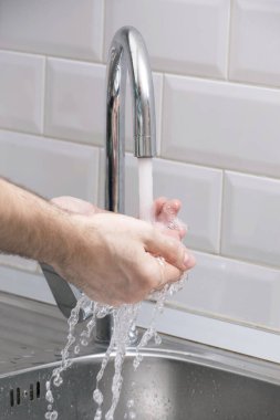 Bir adam ellerini akan suyun altında beyaz bir duvara karşı yıkıyor. Temiz eller kendini virüslerden korur. Ellerimi yıkıyorum..