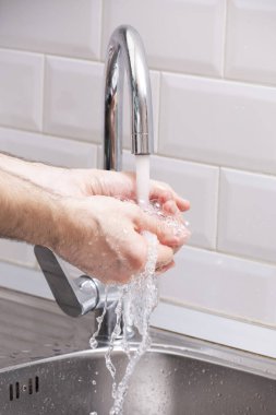 Bir adam ellerini akan suyun altında beyaz bir duvara karşı yıkıyor. Temiz eller kendini virüslerden korur. Ellerimi yıkıyorum..
