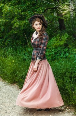 Bahçede yürüyen genç bir kadın. O, eski moda elbise ve peçeli şapka giyiyor. Ağaçlar ve yeşilliklerle çevrili.