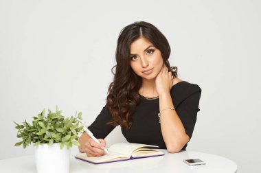 Siyah saçlı, siyah elbiseli, evde çalışan genç bir kız. Masaüstünde bir laptop ve bir saksı çiçek var. İzole edilmiş beyaz arka plan kızı gülümsüyor. İş ona neşe veriyor.