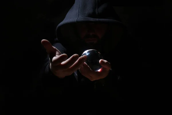 Mann in schwarzer Kapuze mit Kristallkugel beschwört das Böse — Stockbild Mann in schwarzer Kapuze mit Kristallkugel — Stockfoto